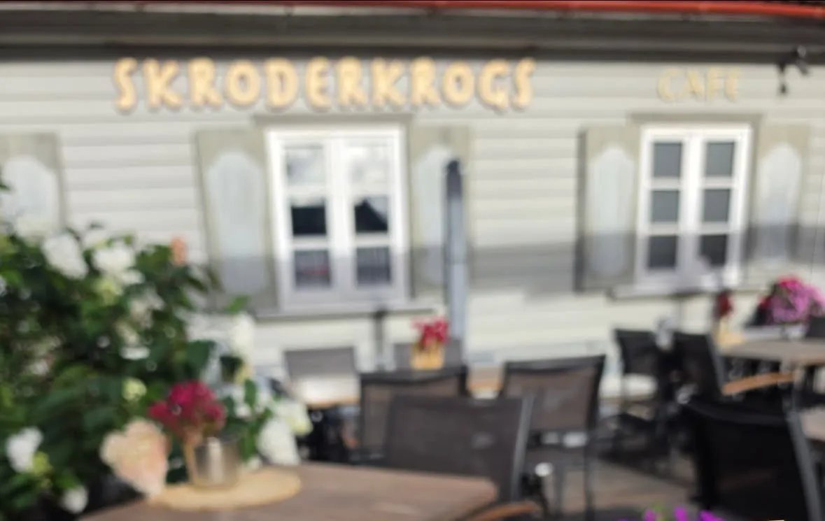 Skroderkroga terase ar galdiem un ziediem, fonā pelēkzaļš koka nams ar uzrakstu „Skroderkrogs”